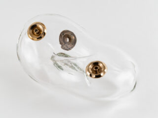 Photograph documenting a transparent glass ring-shaped sculpture with three cast bronze jet nozzles on its surface, against a white background.